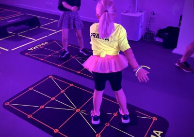 Två barn deltar i en interaktiv träningslek på ett ljust upplyst gym. En flicka i förgrunden i en gul tröja och en rosa kjol hoppar aktivt på en digitalt interaktiv golvyta.