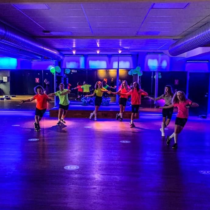 Bild på en gruppdanslektion hos Endorfin, ett träningscenter som finns i Alingsås och Lerum. Deltagarna, klädda i färgglada träningskläder, står i en halvcirkel och utför synkroniserade danssteg. Speglar längs väggen fångar upp deras rörelser och ökar känslan av rymd i det stora, dunkelt belysta rummet. Stämningen verkar energisk och inkluderande, vilket speglar Endorfins fokus på gemenskap och glädje i träningen.