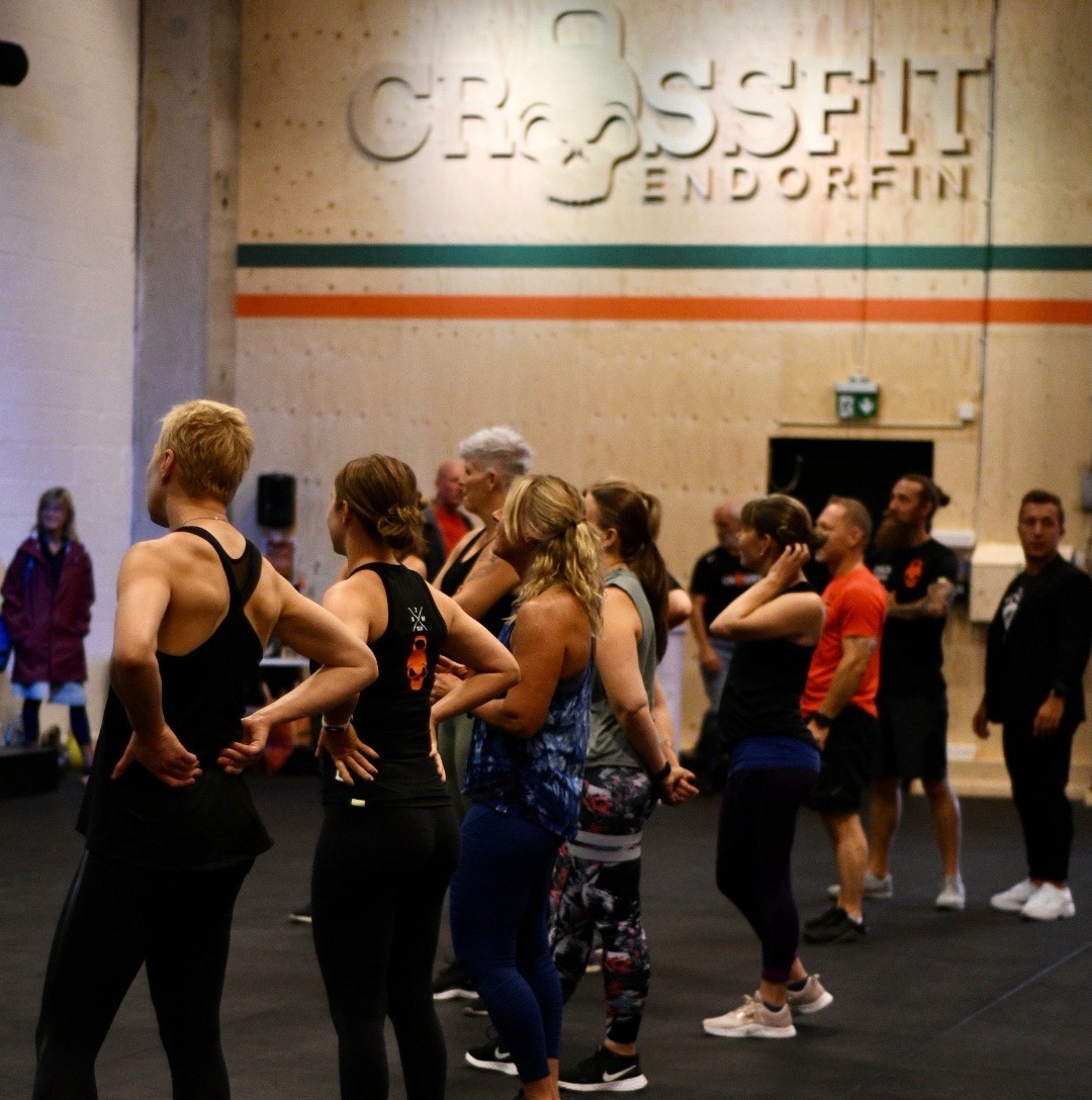 Bild på en gruppträningsklass vid CrossFit Endorfin, med lokaler i både Alingsås och Lerum. Deltagarna, i olika åldrar och klädda i träningskläder, står vända mot instruktören och lyssnar uppmärksamt. I bakgrunden syns CrossFit Endorfins logotyp på väggen, vilket ger en tydlig identitet till lokalen. Atmosfären är energisk och fokuserad, med deltagare som är redo att börja träningen.