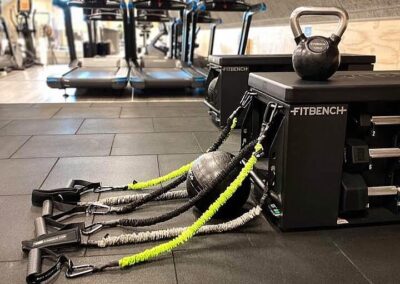 Närbild av en svart "FITBENCH" med flera TRX-band och en kettlebell på gymmets golv, vilket indikerar en mångsidig träningsstation.